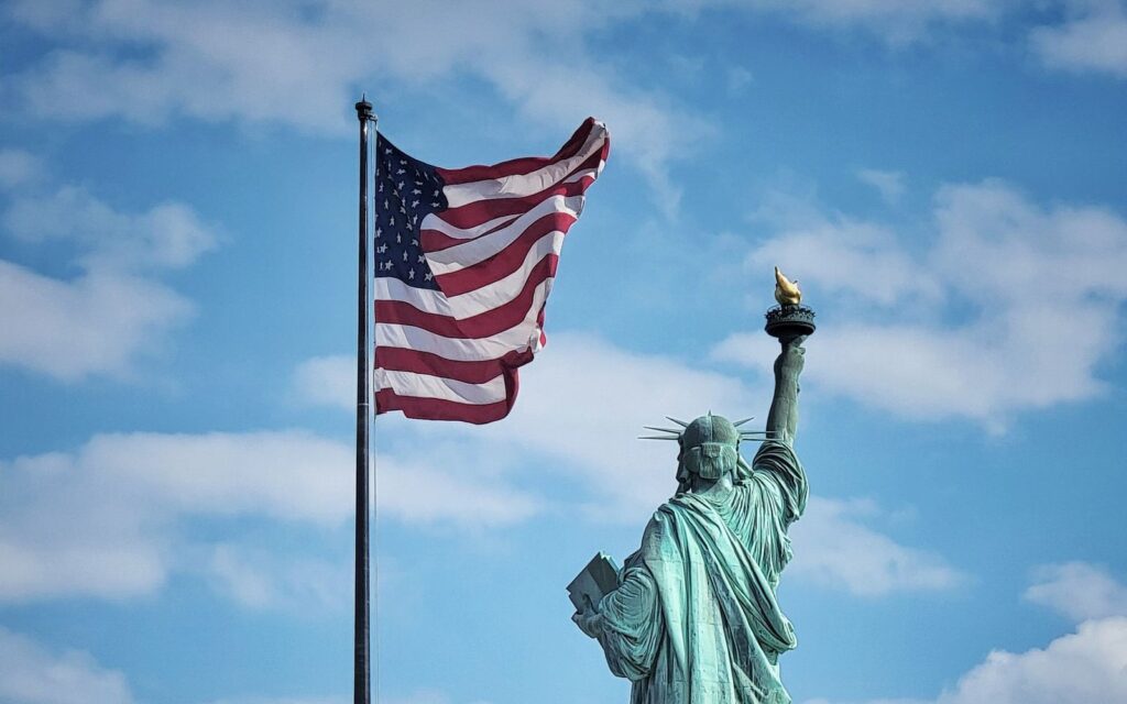 Statue of Liberty beside a waving American flag against a blue sky